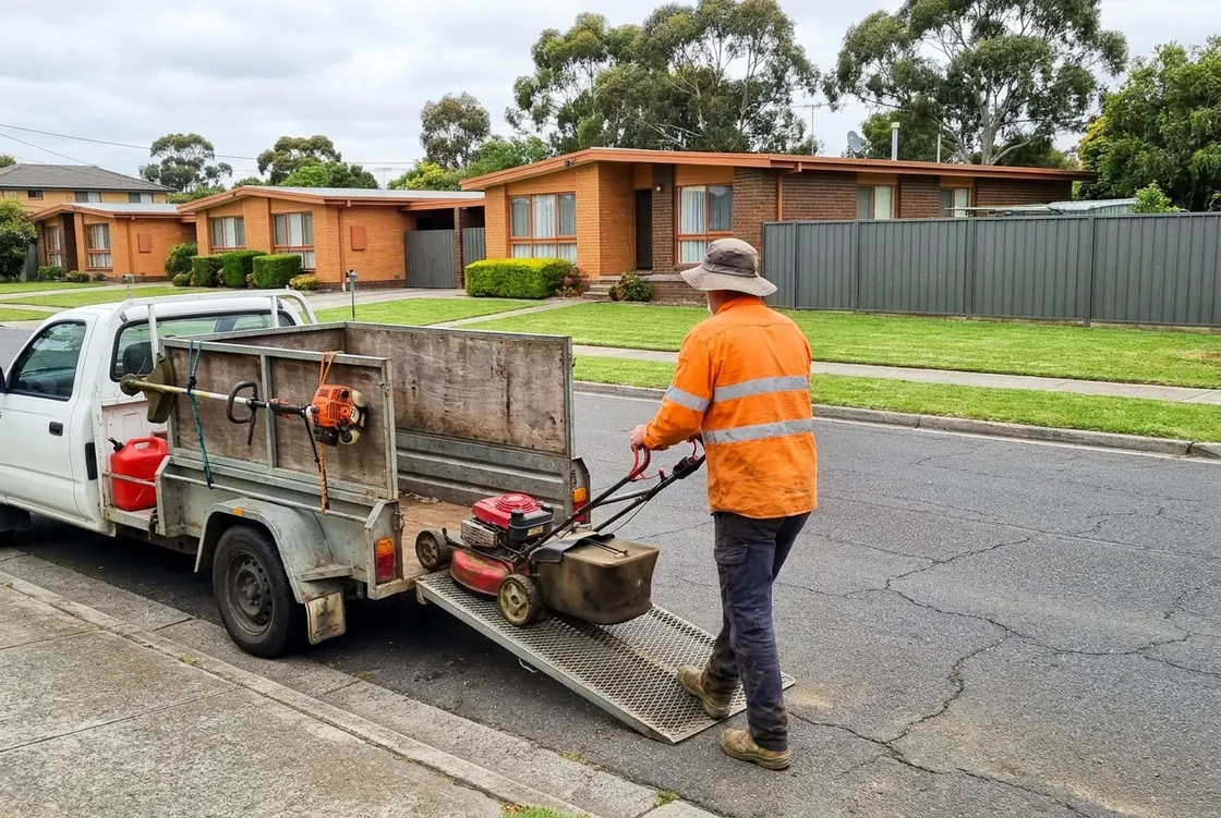 A mowing contractor seen from behind in a hi-vis shirt and wide-brim hat wheels a petrol mower up the ramp of a box trailer hitched to a white ute, parked outside a row of brick units with freshly mown lawns in Melbourne's north